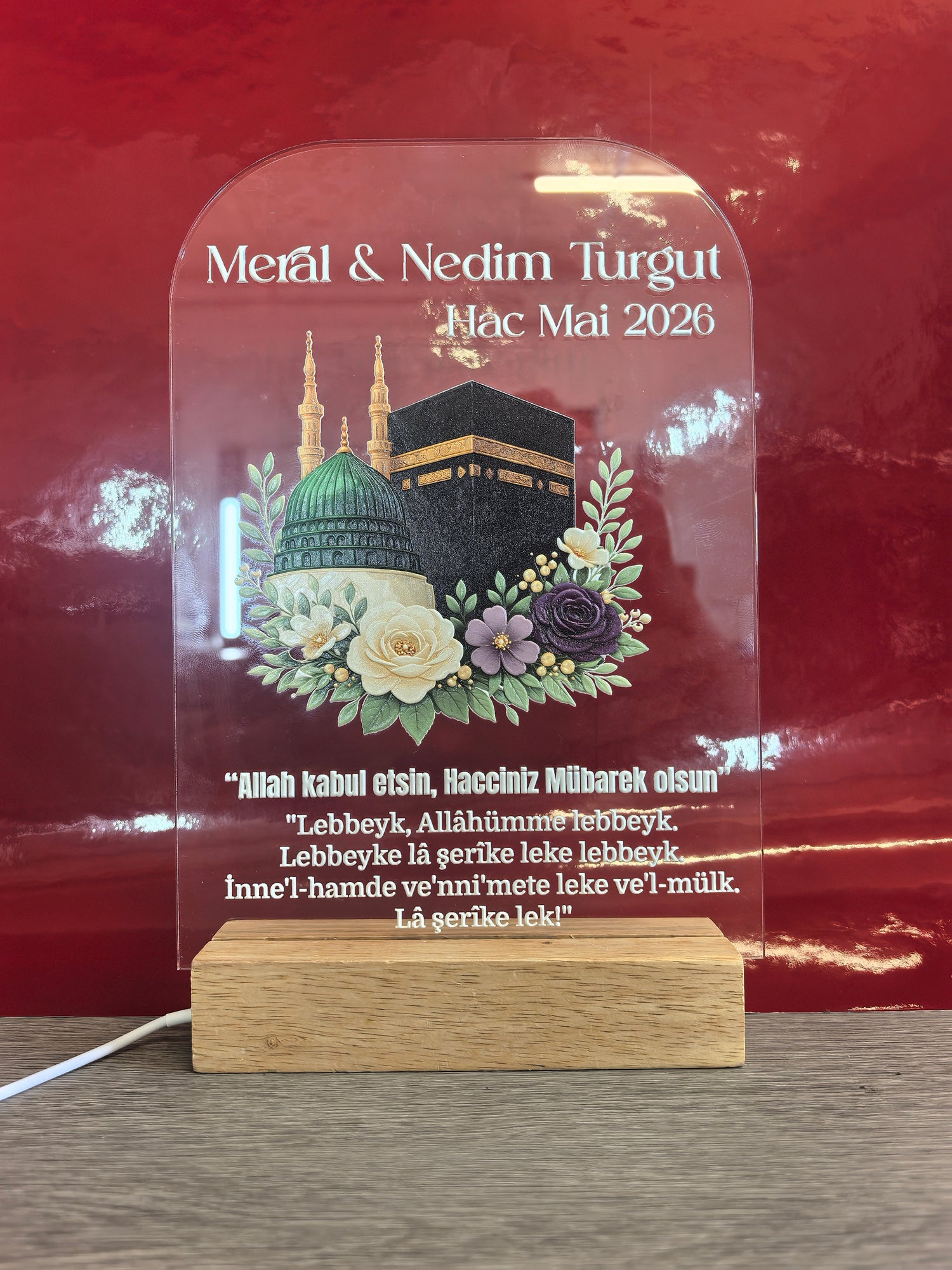 Personalisiertes Hac Acrylglas mit LED-Holzsockel, Kaaba und Moschee-Motiv mit Blumendekoration, gravierte Namen Meral & Nedim Turgut, Hac Mai 2026 und Talbiyya-Gebet, roter Hintergrund